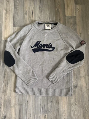 Grå stickad tröja från Morris - Snygg grå stickad tröja från Morris med mörkblå elbow patches och broderad logga på bröstet. Tröjan har ribbade muddar och nederkant samt en patch med brittiska flaggan på ärmen. Perfekt för dig som gillar klassisk men trendig stil.