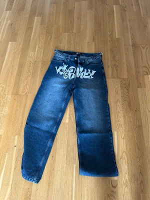 Blå jeans med graffiti från Vicinity - Säljer ett par blå jeans från Vicinity i storlek M med bred passform och coolt graffiti-tryck i vitt över framsidan. Jeansen har klassisk femficksdesign, bälteshällor och dragkedjegylf. Perfekta för dig som gillar streetwear och vill sticka ut.