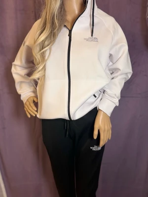 Vit hoodie från The North Face - Snygg vit hoodie från The North Face med dragkedja framtill och svart logga på bröstet. Hoodien har huva med justerbara snören och ribbade muddar vid ärmsluten. Perfekt för en chill och sportig look. Materialet känns mjukt och bekvämt.