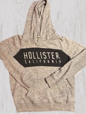 Grå Hollister hoodie med tryck - Snygg gråmelerad hoodie från Hollister med stor svart logga och texten 'Hollister California' på bröstet. Hoodien har huva med dragsko och ribbade muddar vid ärmar och nederkant. Perfekt för en avslappnad och trendig look.