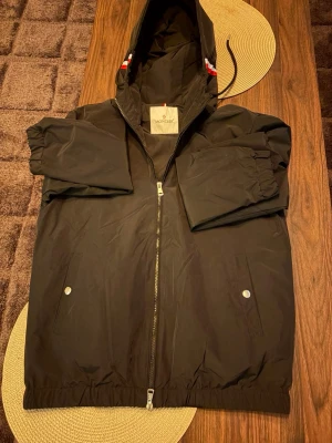 Svart vindjacka från Moncler - Snygg svart vindjacka från Moncler med huva och dragkedja framtill. Jackan har två fickor med tryckknappar och elastiska muddar vid ärmsluten. Detaljer med Monclers logga och diskreta rödvita ränder på huvan. Perfekt för blåsiga dagar.