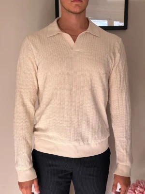 Beige långärmad pikétröja från Riley - Stilren beige långärmad pikétröja i storlek L med diskret ribbad struktur och klassisk krage. Tröjan har en relaxed fit och breda muddar vid ärmslut och midja. Perfekt för en clean och avslappnad look.