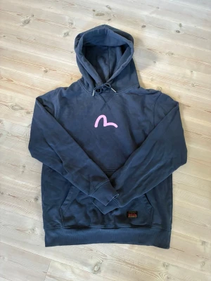 Blå hoodie från Evisu med rosa tryck - Säljer en blå hoodie från Evisu med en rosa logga på bröstet. Skitfet och i bra skick, nypris på över 1500.