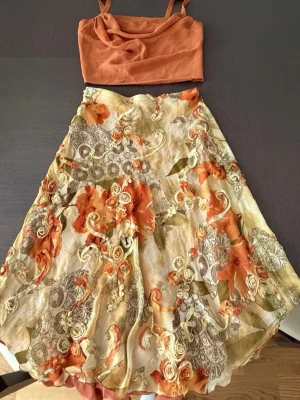 Blommig långkjol i orange och beige - Säljer en unik långkjol med blommigt mönster i orange, beige och grönt plus en matchande topp till. Kjolen har volangdetaljer och ett skirt, lätt material som ger en flowy känsla. Perfekt för dig som vill sticka ut med en bohemisk och färgstark stil.