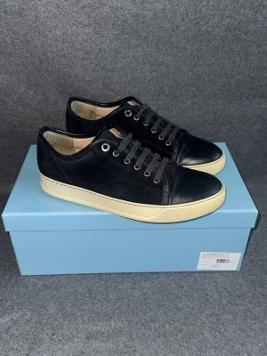 Svarta sneakers med vit sula från Lanvin - Stilrena svarta sneakers med ovandel i mocka och läderdetaljer från Lanvin. Skorna har vita, tjocka gummisulor och svarta skosnören. Klassisk låg modell med metallöljetter och diskret design som passar till det mesta.