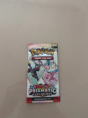 Pokémon Prismatic Evolutions booster - Ett oöppnat Pokémon booster pack från Scarlet & Violet Prismatic Evolutions. Förpackningen är färgglad med illustrationer av olika Pokémon och innehåller 10 samlarkort. Perfekt för dig som gillar att samla eller spela Pokémon Trading Card Game.