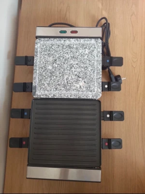 Raclettegrill med sten- och grillplatta - Elektrisk raclettegrill med både en grå stenplatta och en svart grillplatta. Plats för åtta små pannor med handtag, perfekt för att laga mat tillsammans. Utrustad med strömkabel och indikatorlampor. Passar för hemmakvällar och middagar med vänner.