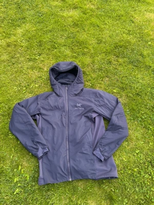 Mörkgrå vindjacka från Arc’teryx - Snygg mörkgrå vindjacka från Arc'teryx med huva och hel dragkedja framtill. Jackan har minimalistisk design, diskret logga på bröstet och är tillverkad i lätt, vindtätt material som passar perfekt för utomhusaktiviteter.