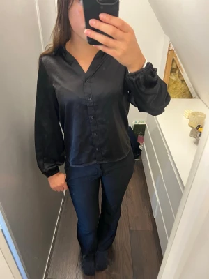 Svart satinblus från Vero Moda - Stilren svart blus i glansigt satinmaterial från Vero Moda. Blusen har V-ringning, knappar framtill och långa ärmar med resår vid handlederna. Aldrig använd
