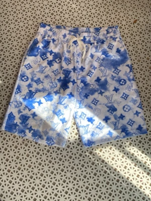 Lv shorts  - Säljer ett par blåvita shorts med lv mönster och snörning i midjan. Shortsen har ett unikt batikliknande tryck med olika symboler och elastisk midja för skön passform. Perfekta för sommardagar eller strandhäng.