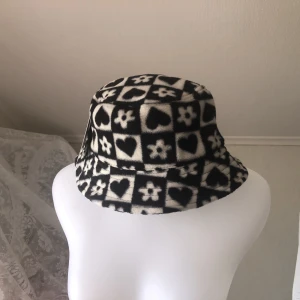 Buckethat - Fluffig buckethat i ”rutigt schack mönster” i svart och vitt med hjärtan och blommor på! Från H&M divided i nyskick prislapp sitter kvar men köpt för nåt år sen så finns inte kvar på H&M. Super mysig å ha:) Läs gärna bio<3
