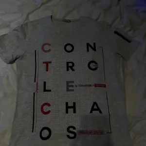 Ljusgrå tunn skön tshirt med svart-rött tryck ”Controle Chaos”. Liten dragkedja på ena armen. Storlek L. Knappt använd