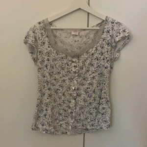 Mönstrat T-shirt  - Vit top med blå blommor som mönster. Vit spets vid kragen. Köpt andra hand men aldrig använd av säljaren. Ok skick. 