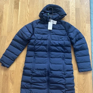 Oanvänd Dunjacka från Henry Lloyd stl M dam  - Längre dunjacka med ett vind- och vattenavvisande material och en normal passform. Avtagbar huva med justerbar dragsko och hög krage. Två fickor fram och justerbar dragsko i midjan. Färg marinblå. Storlek M  Helt oanvänd.