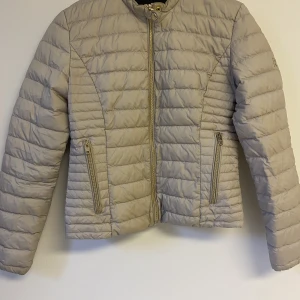 Dunjacka rockandblue - Beige dunjacka med guld detaljer från rockandblue. Perfekt till hösten då den inte är jättetjock. Använd ett fåtal gånger, säljer pga för liten. Nypris 1600kr
