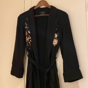 Kavaj/kaftan - Svart kaftan med blommor och med knyt i midjan. Fickor framtill. XS/S men passar upp till M