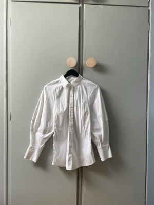 COS skjorta med ballongärmar och markerad midja strl S - Fin skjorta med ballongärmar och markerad midja. I mycket gott skick! I strl S. Helt slutsåld på hemsidan.   Material 64% Bomull, 5% Elastan, 31% Polyamid  Färg Vit