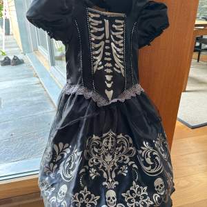 Svart Halloween klänning med silvrigt tryck; skelett på bröstet, döskallar m.m. Underkjolen står ut med hjälp av ställning. Använd en gång. 