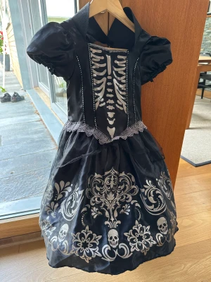Halloween klänning stl 116 (6-7 år) - Svart Halloween klänning med silvrigt tryck; skelett på bröstet, döskallar m.m. Underkjolen står ut med hjälp av ställning. Använd en gång. 