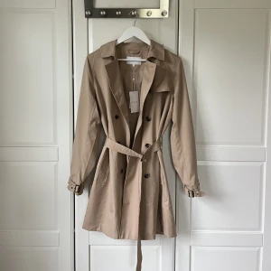 Trenchcoat - Superfin oanvänd trenchcoat från Vila. Prislapp kvar (ordinarie pris 599 kr). Storlek 38, passar den som vanligen har S i storlek.