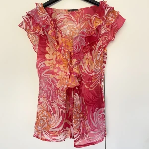 Blommig blus med volanger - Ärmlängd 14, bredd 41, längd 59. Blus i orange, rosa, vinrött och vitt. Färgerna är mer vibranta än vad bilderna visar! Tunt och svalt material, v-ringning, volanger vid ringning och ärmar och blixtlås i vänster sida💖💖 Köp via ”Köp Nu”💫