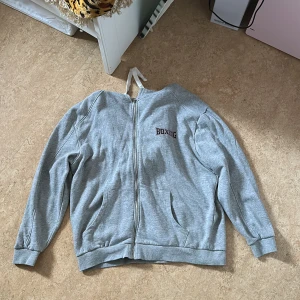 Grå zip hoodie - En grå zip Hood som är lite oversized vid armarna men annars sitter väldigt bra. Väljer att sälja eftersom den inte kommer att komma till användning längre då trycket inte var så förtjusande helt enkelt.