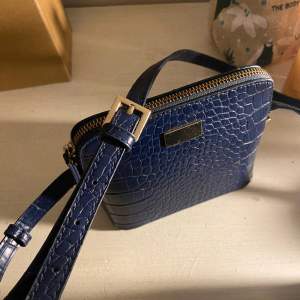 Säljer denna krokodilmönstrade marinblå väska från Åhléns. Väskan är i bra och fräscht skick, och är endast använd ett fåtal gånger. Bandet är lite längre men går att korta ned om man skulle vilja det.