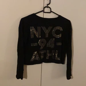 Svart tröja med guld text - Säljer denna snygga svarta tröjan med guld ”NYC” text, då den blivit för liten. Använd fåtal gånger och är i bra skick! Det står ingen storlek på tröjan men den är som en xxs-xs