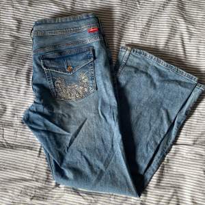 Ett par jeans i lätt stretchig bomullsdenim. Jeansen har låg midja och vida ben med utsvängda benslut. Gylf med dragkedja och tryckknappar. Framfickor, en myntficka och bakfickor med lock och tryckknapp. Jeansen är knappt använda.