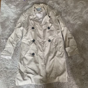 Trenchcoat - Stilren kappa perfekt till hösten. Saknas en knapp men kan lätt sys på då extra knappar finns. Band i midjan ingår 