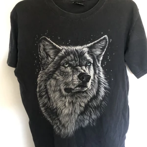 T shirt med tryck i stl L. - T shirt med tryck. T shirten är i en urtvättad svart färg. Trycket är en varg med blåa ögon. T shirten är i fint skick och i stl L och gjord av bomull.