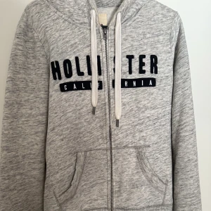 Hollister zip hoodie - Ziphoodie från Hollister. Ljusgrå med svart text. Har plats för tummarna fram vid muddarna. Sparsamt använd