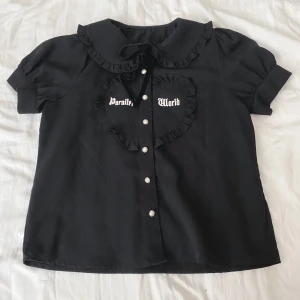 Ank Rouge - Blus - Blus från Ank Rouge med pärlknappar, hjärta och ”parallel world” tryck. Blusen kommer med ett sammetsband för kragen och är i gott skick. Säljer då den ej kommer till användning. Köptes från deras hemsida förra året och endast använts fåtal gånger sen des