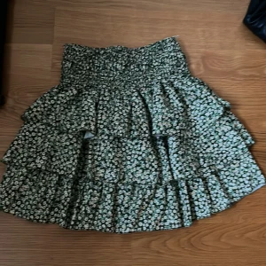 Blommig kjol med volanger - Säljer en superfin blommig kjol i grönt med små vita och gula blommor. Kjolen har flera lager med volanger som ger en härlig volym och en elastisk midja för extra komfort. Perfekt för sommarens alla fester eller en dag på stan!