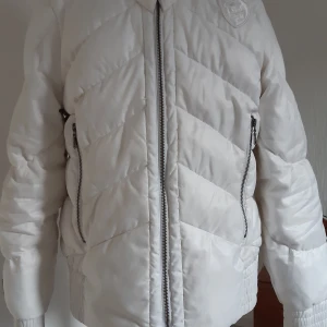 Vit pufferjacka med dragkedja - Säljer en snygg vit pufferjacka med coola detaljer. Jackan har en dragkedja framtill och två dragkedjefickor på sidorna. Den är quiltad och har ett stilrent märke på bröstet. Perfekt för vintern och håller dig varm och trendig!