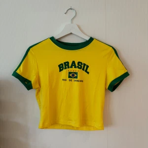 Gul Brasil T-shirt - Säljer en  gul T-shirt med texten 'BRASIL' och 'RIO DE JANEIRO' samt en brasiliansk flagga på framsidan. Den har gröna detaljer vid krage och ärmslut, och är kortärmad. 