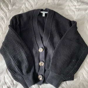 Svart stickad kofta från H&M - Säljer en snygg svart stickad kofta från H&M. Den har en klassisk design med stora knappar framtill och ribbade detaljer. Perfekt för höst och vinter, och den är mysig och bekväm att ha på sig. Passar både till vardags och lite finare tillfällen.