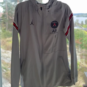 Grå vindjacka från Jordan x PSG - Säljer en grå vindjacka från Jordan i samarbete med Paris Saint-Germain. Jackan har en cool design med PSG-loggan och Jordan-märket på bröstet. Den har långa ärmar med röda och svarta detaljer på ärmarna. Perfekt för sportiga tillfällen eller som en stilren vardagsjacka.