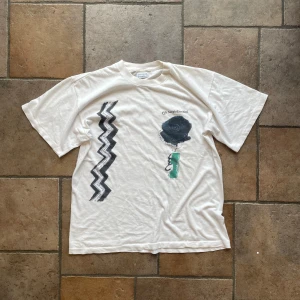 Vit t-shirt från Sergio Tacchini - Säljer en cool vit t-shirt från Sergio Tacchini. Den har ett unikt tryck med en svart spiralformad design och en grön detalj på framsidan. T-shirten är kortärmad och i normal passform, perfekt för en avslappnad stil. Tillverkad i Italien