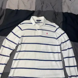 Säljer en snygg vit och blå randig pikétröja från Ralph Lauren. Den har långa ärmar och en klassisk krage. Tröjan är i nyskick och har det ikoniska röda broderade logotypen på bröstet. Perfekt för både vardag och lite finare tillfällen!