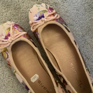 Blommiga ballerinaskor från Tamaris - Säljer ett par supergulliga ballerinaskor från Tamaris. De är i en ljusrosa nyans med ett vackert blommigt mönster i olika färger. Skorna har en söt rosett framtill! De är i storlek 37 och i nyskick.💞