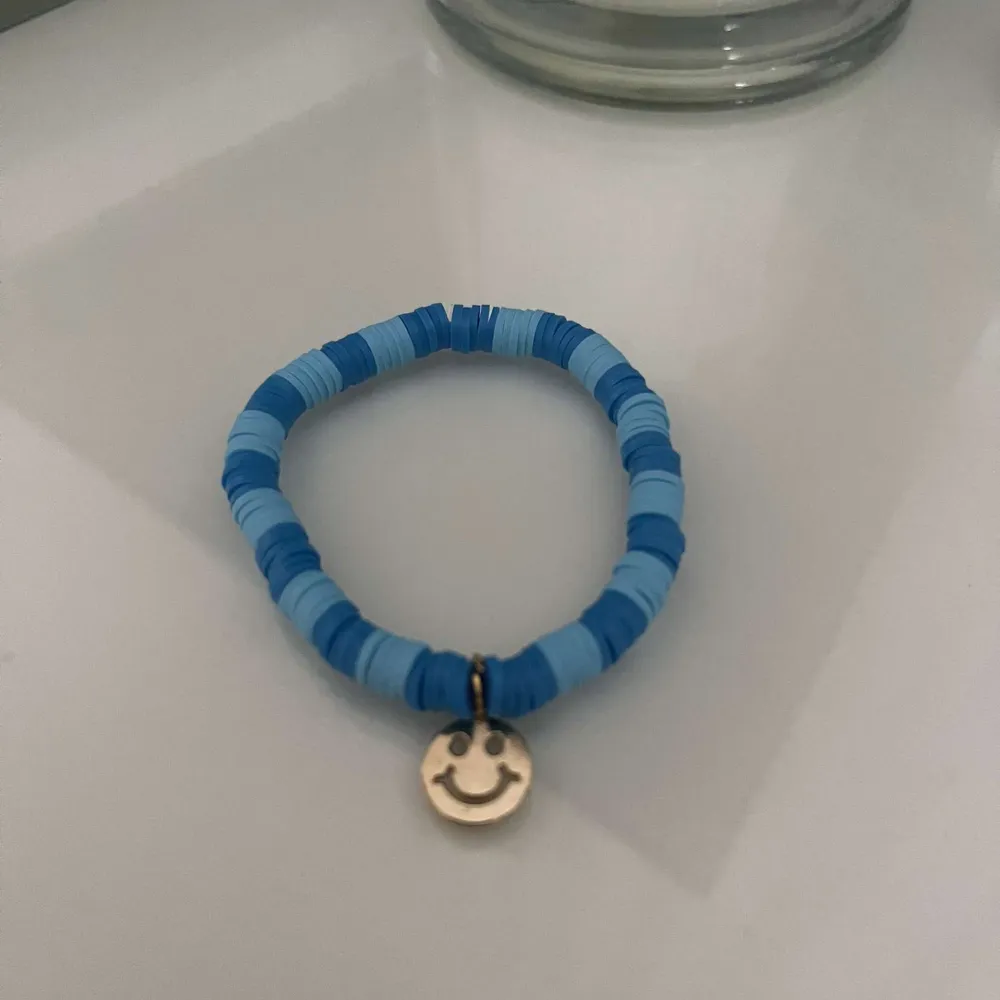 Säljer ett supergulligt armband i olika nyanser av blått. Det har en liten guldberlock med en glad smiley på. Perfekt för att liva upp vilken outfit som helst! 💙. Accessoarer.