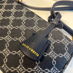 Holly & Whyte axelväska - Passar utmärkt till en festligare tillställning. Marin blå och vit mönster med guld-detaljer. Väskan rymmer mer än en mobil, trådlösa hörlurar m.m