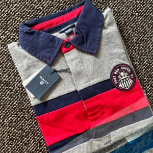 Randig pikétröja från GANT - Säljer en snygg och stilren randig pikétröja från GANT till barn. Tröjan har en klassisk krage i mörkblått med knappar och ett coolt märke på bröstet. Perfekt för både vardag och lite finare tillfällen!