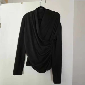 Svart omlottopp - Säljer en elegant svart omlottopp med långa ärmar. Perfekt för höst och vinter, den har en draperad design som ger en snygg och stilren look. Passar både till vardags och festliga tillfällen. Passar s/m