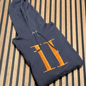 Mörkblå hoodie med orange tryck - Säljer en snygg mörkblå hoodie från LES DEUX. Den har en klassisk känguruficka och justerbar huva med dragsko. Perfekt för en avslappnad stil!