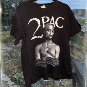 Svart 2Pac t-shirt från Fruit of the Loom - Säljer en svart t-shirt med ett stort tryck av 2Pac på framsidan. T-shirten är från Fruit of the Loom och är i storlek M. Perfekt för dig som gillar klassisk hiphop-stil. Den är kortärmad och gjord i bomull, vilket gör den bekväm att bära. Passar bra till vardags eller för en avslappnad look.