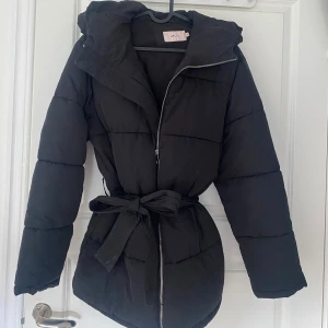 Svart pufferjacka med bälte - Säljer en snygg svart pufferjacka som är perfekt för kalla dagar. Den har en dragkedja framtill och ett bälte i midjan för en fin passform. Jackan är långärmad och supermysig att ha på sig. Perfekt för både höst och vinter!