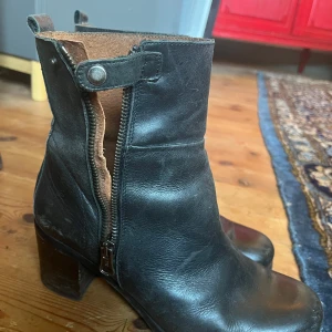 Svarta skinnstövletter med dragkedja - Säljer ett par svarta skinnstövletter i välanvänt skick. De har en praktisk dragkedja på sidan och en stabil klack som ger lite extra höjd. Perfekta för höst och vinter, och de är tillverkade i Spanien. Passar bra till både vardag och fest.