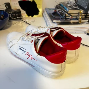 Vita sneakers från Tommy Hilfiger - Säljer ett par snygga vita sneakers från Tommy Hilfiger. De har en stilren design med röd insida och detaljer. Skorna har snörning och är i nyskick. Perfekta för både vardag och fest!